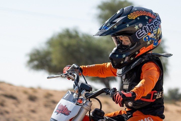 Child in orange gear riding a dirt bike, wearing a helmet with stars and text, desert background.