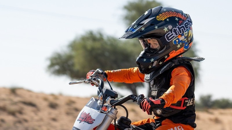 Child in orange gear riding a dirt bike, wearing a helmet with stars and text, desert background.