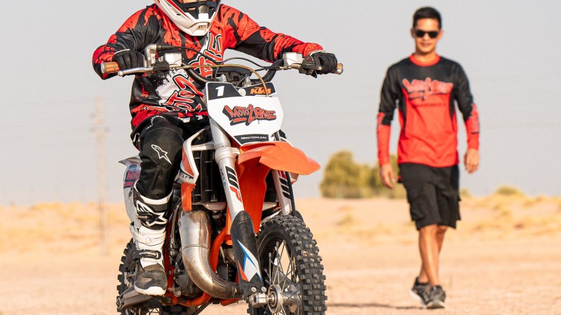 Child riding a dirt bike on a sandy path with an adult walking behind.