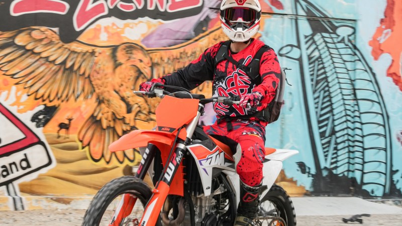 Rider on an orange dirt bike wearing red gear in front of a colorful mural.