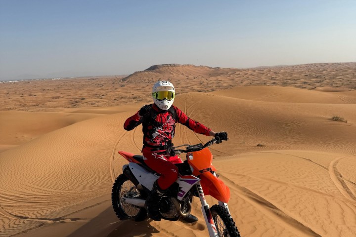 Motorcyclist in red gear on orange dirt bike atop desert sand dunes.