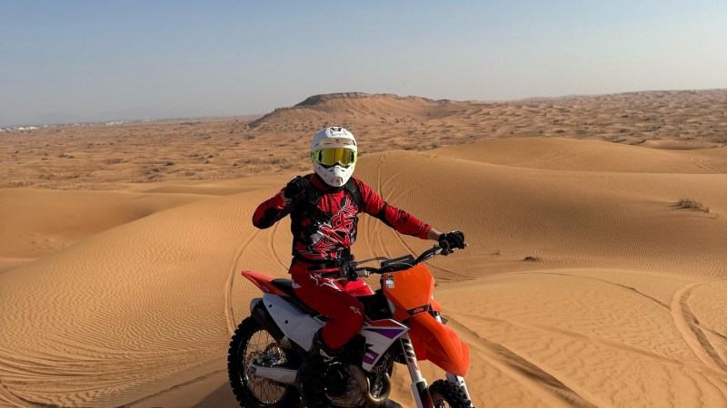 Motorcyclist in red gear on orange dirt bike atop desert sand dunes.