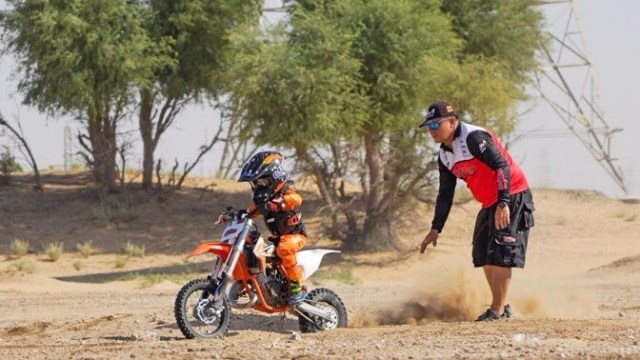 Child on dirt bike with adult supervising in a desert landscape.