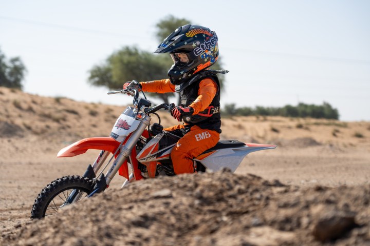Child in orange gear riding a dirt bike on a sandy track with trees in the background.