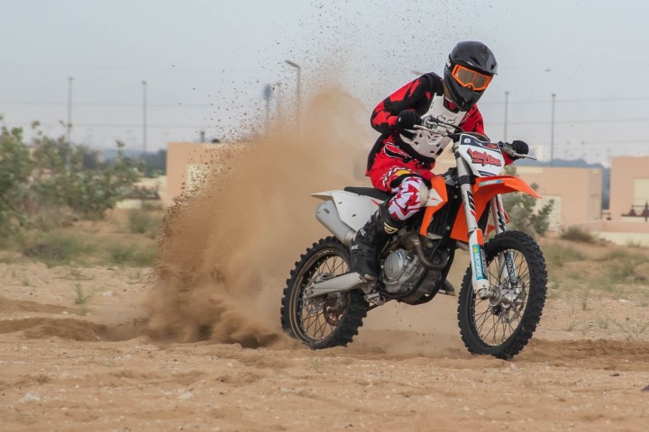 Person on a dirt bike kicking up sand on a track, wearing red and black gear.