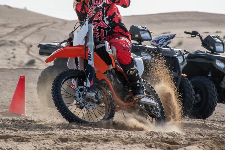 Person on a dirt bike turning in sand near an orange cone, dressed in red and black gear.