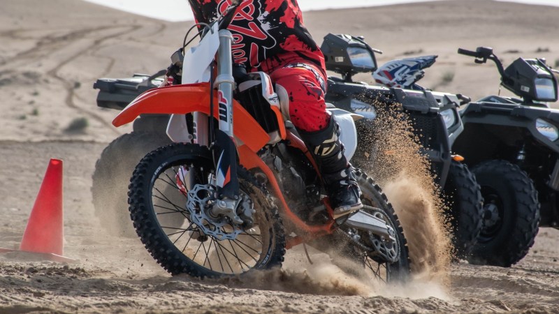 Person on a dirt bike turning in sand near an orange cone, dressed in red and black gear.