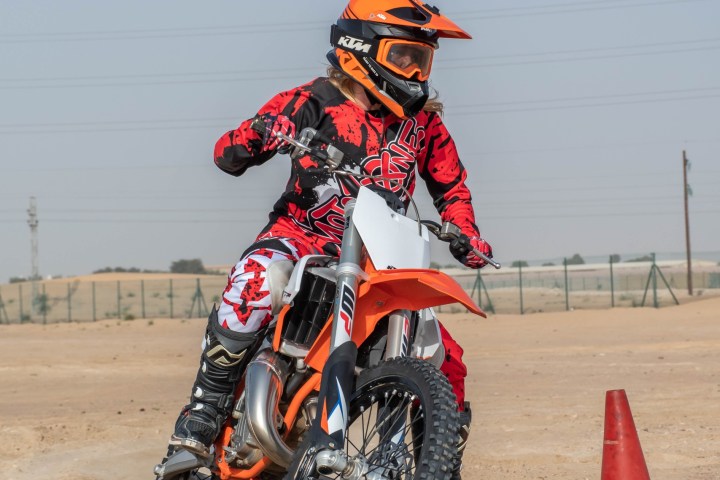 Person in orange and black gear riding a dirt bike on sandy terrain near a red cone.