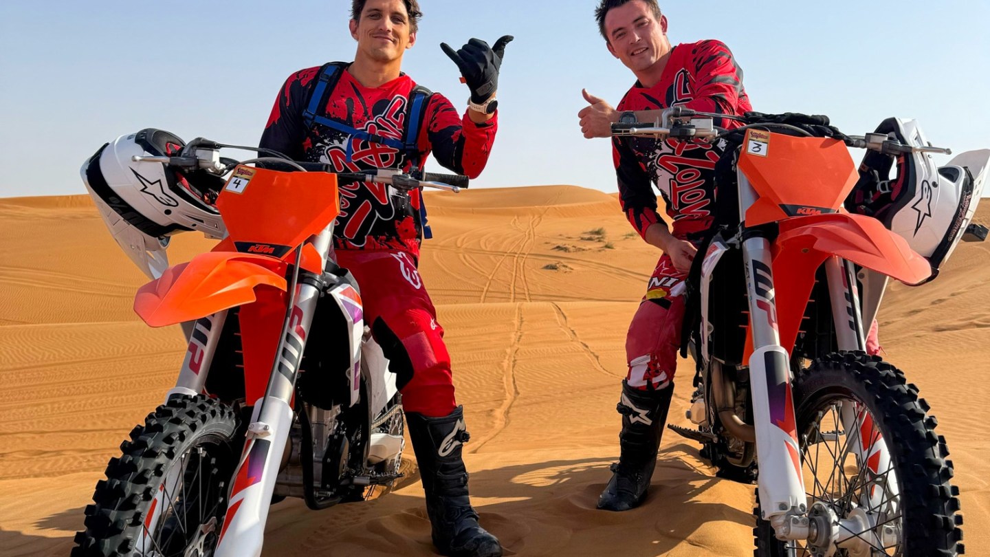 Two people in motocross gear posing with dirt bikes on sand dunes.