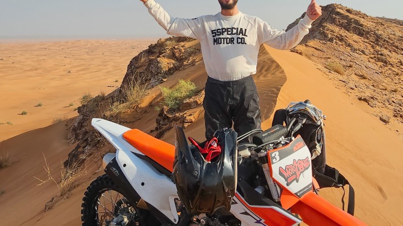 Person on desert sand dune with a dirt bike, arms raised, blue sky background.