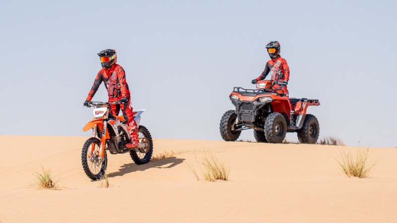 Two riders in red gear on a dirt bike and ATV in a sandy desert landscape.