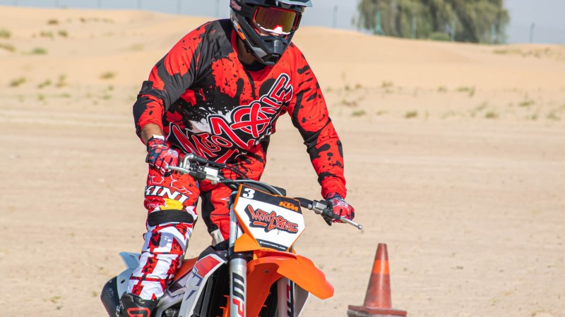Person in red gear riding an orange dirt bike on sandy terrain with a traffic cone nearby.