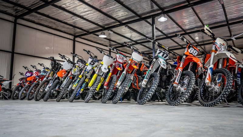 Row of colorful dirt bikes in a garage with metal ceiling and bright lighting.