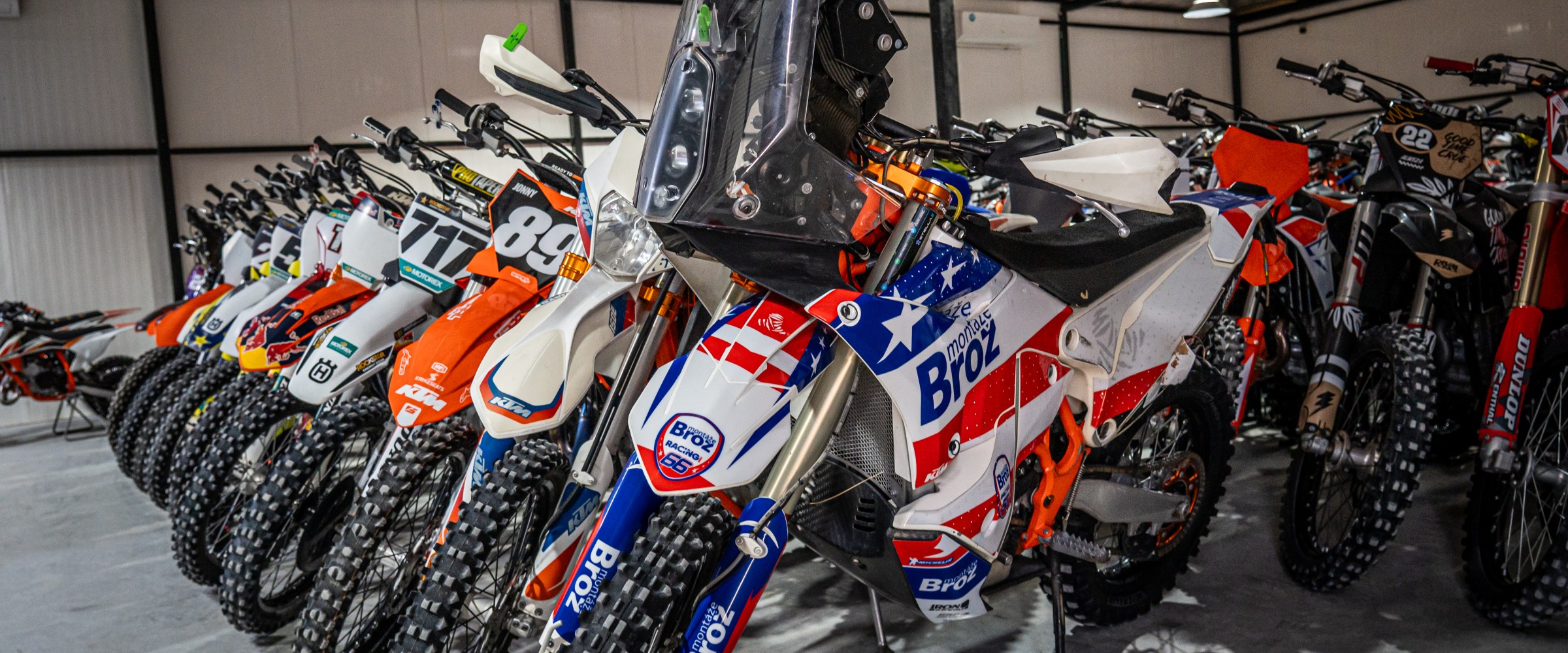 Line of dirt bikes in a warehouse, each with unique decals and numbers, displayed in rows under a metal roof.