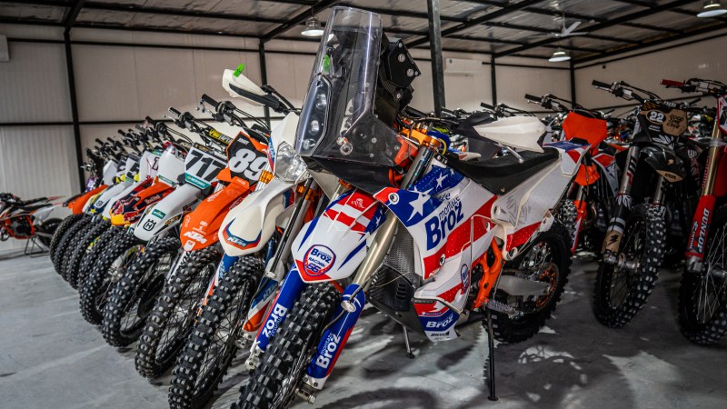 Line of dirt bikes in a warehouse, each with unique decals and numbers, displayed in rows under a metal roof.