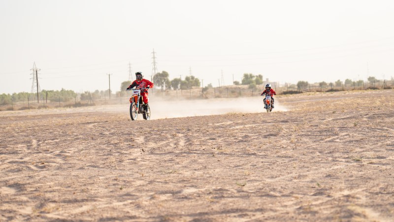 Two motocross riders on dirt track, kicking up dust under a clear sky.