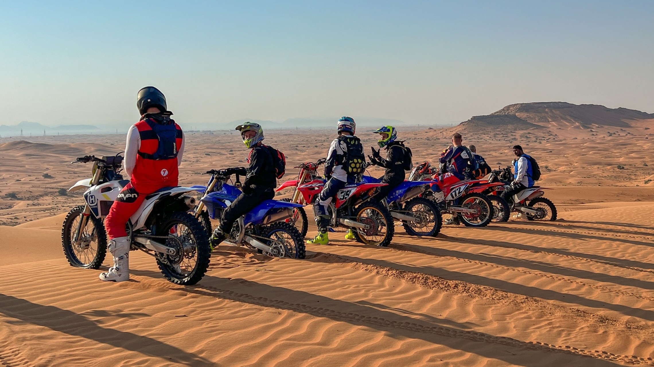 Group of motocross riders on bikes atop sand dunes, desert landscape.
