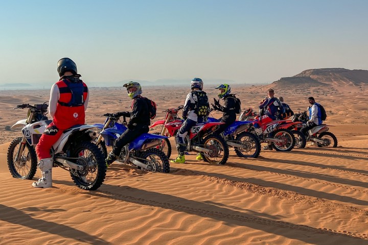 Group of motocross riders on bikes atop sand dunes, desert landscape.