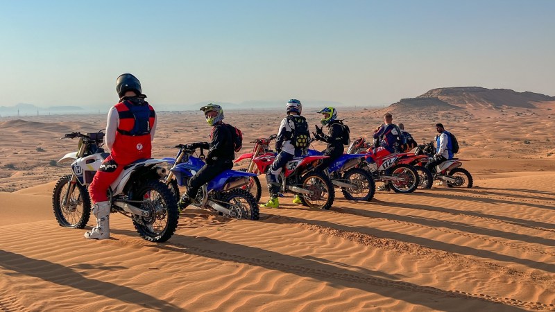 Group of motocross riders on bikes atop sand dunes, desert landscape.