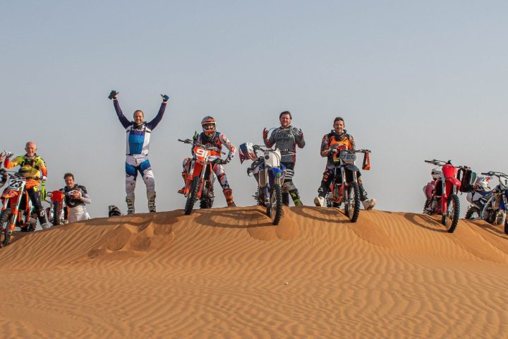 Group of motocross riders posing with bikes on a desert dune.