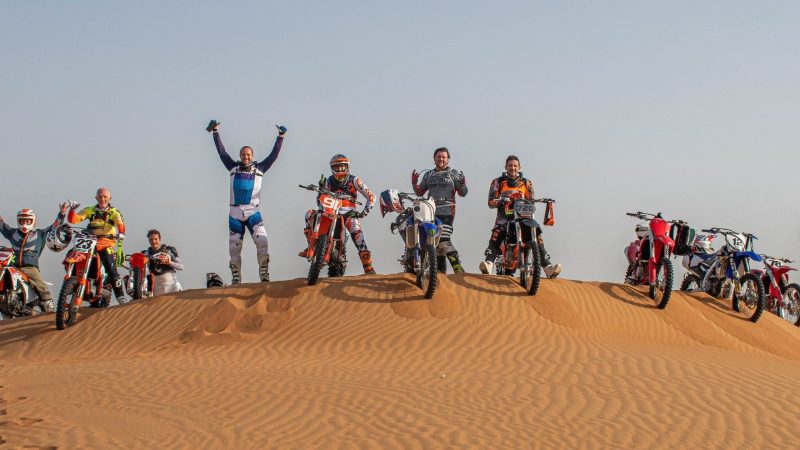 Group of motocross riders posing with bikes on a desert dune.