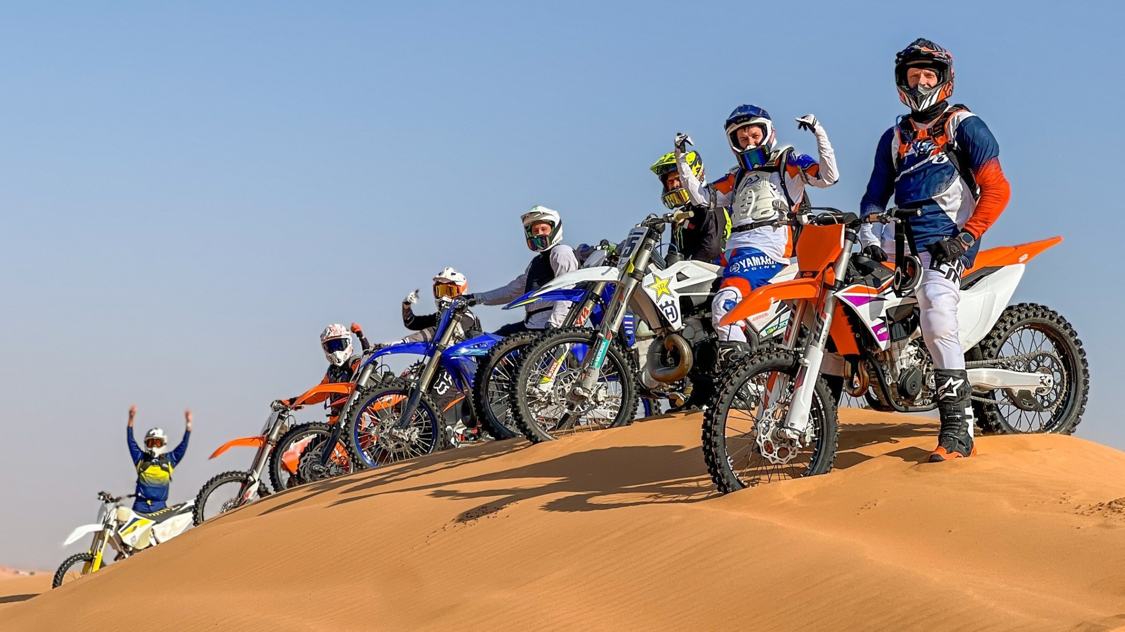 Group of motocross riders posing on dirt bikes on a sand dune.