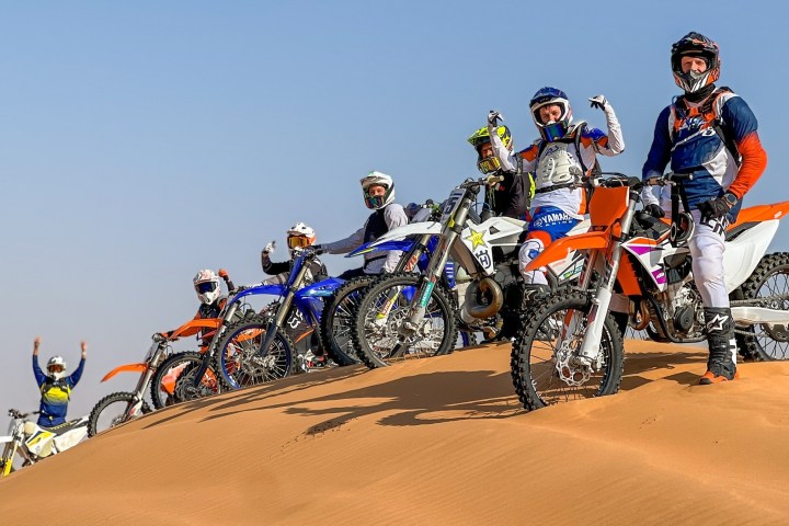 Group of motocross riders posing on dirt bikes on a sand dune.