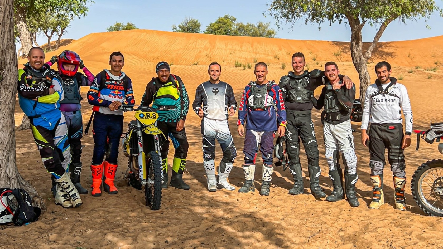 Group of motorcyclists in gear standing by dirt bikes with desert background.