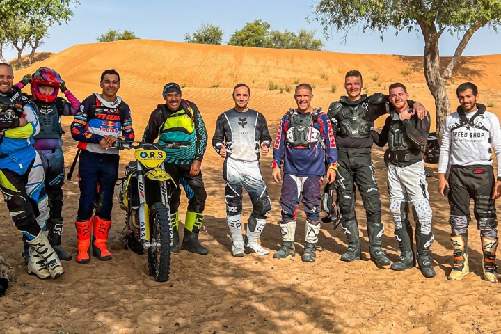 Group of motorcyclists in gear standing by dirt bikes with desert background.