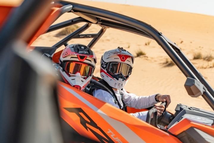 Two people in helmets driving an orange dune buggy in the desert.