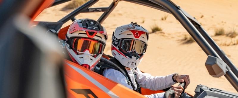 Two people in helmets driving an orange dune buggy in the desert.
