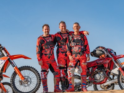 Three men in red motocross gear stand between two dirt bikes on sand under a clear blue sky.