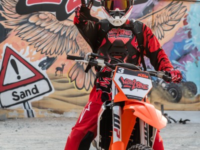 Motorcyclist in red gear poses with a motorbike, colorful graffiti background.
