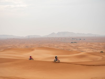 Two people riding dirt bikes on sand dunes under a cloudy sky.