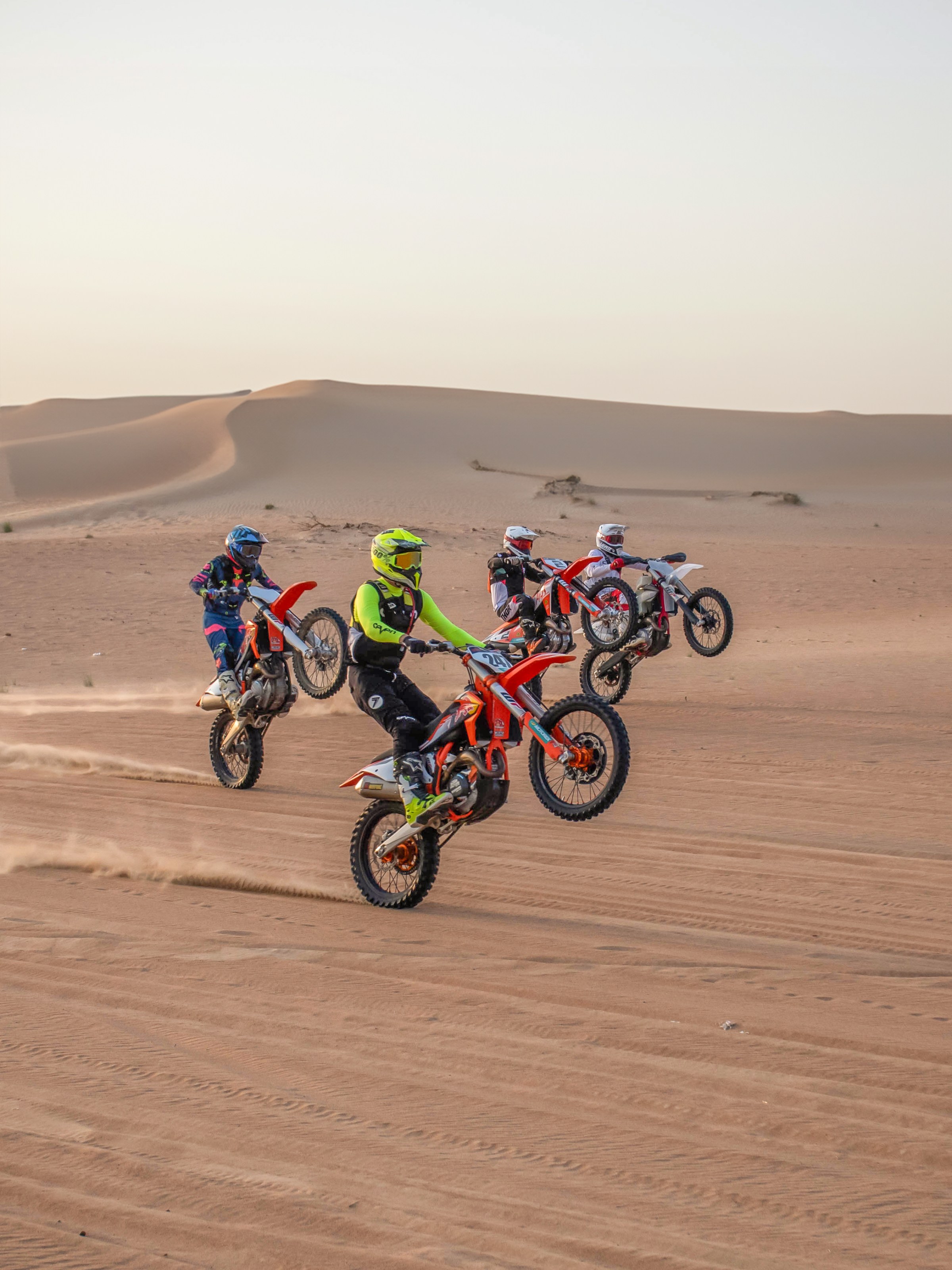 Four motorcyclists doing wheelies on dirt bikes in desert terrain with sand dunes in background.