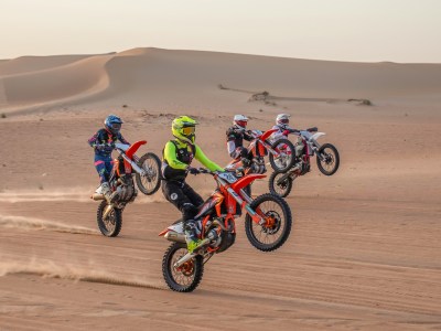 Four motorcyclists doing wheelies on dirt bikes in desert terrain with sand dunes in background.