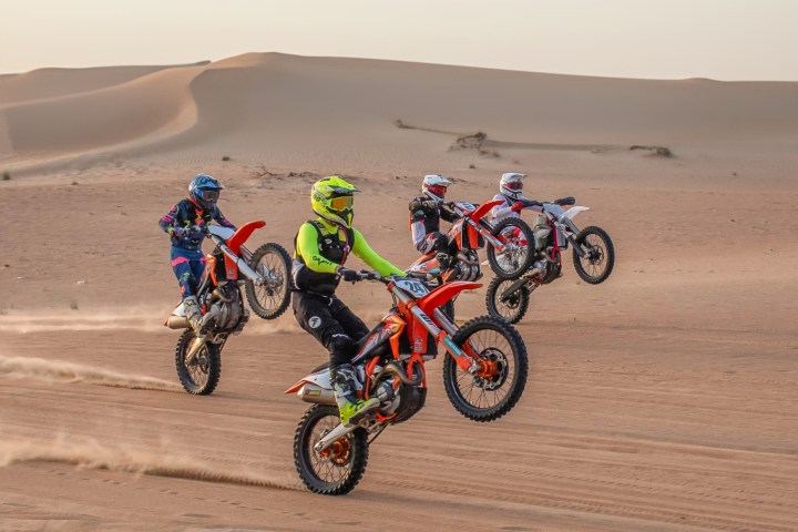 Four motorcyclists doing wheelies on dirt bikes in desert terrain with sand dunes in background.