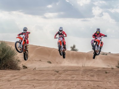 Three motorcyclists jumping over sand dunes under a cloudy sky.
