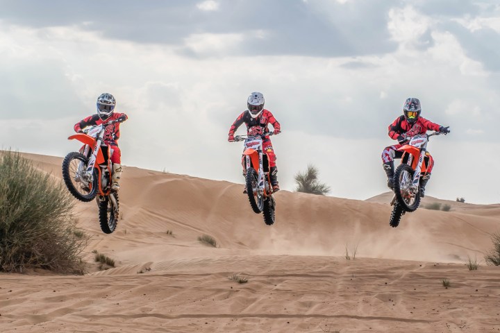 Three motorcyclists jumping over sand dunes under a cloudy sky.