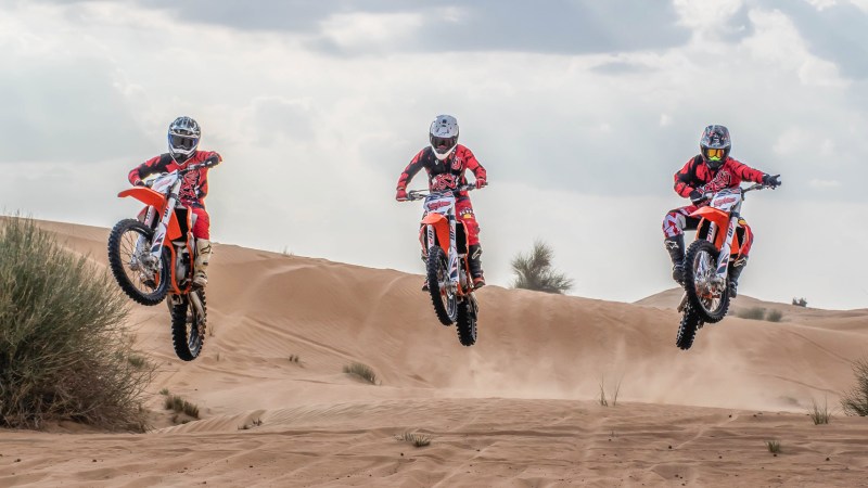 Three motorcyclists jumping over sand dunes under a cloudy sky.