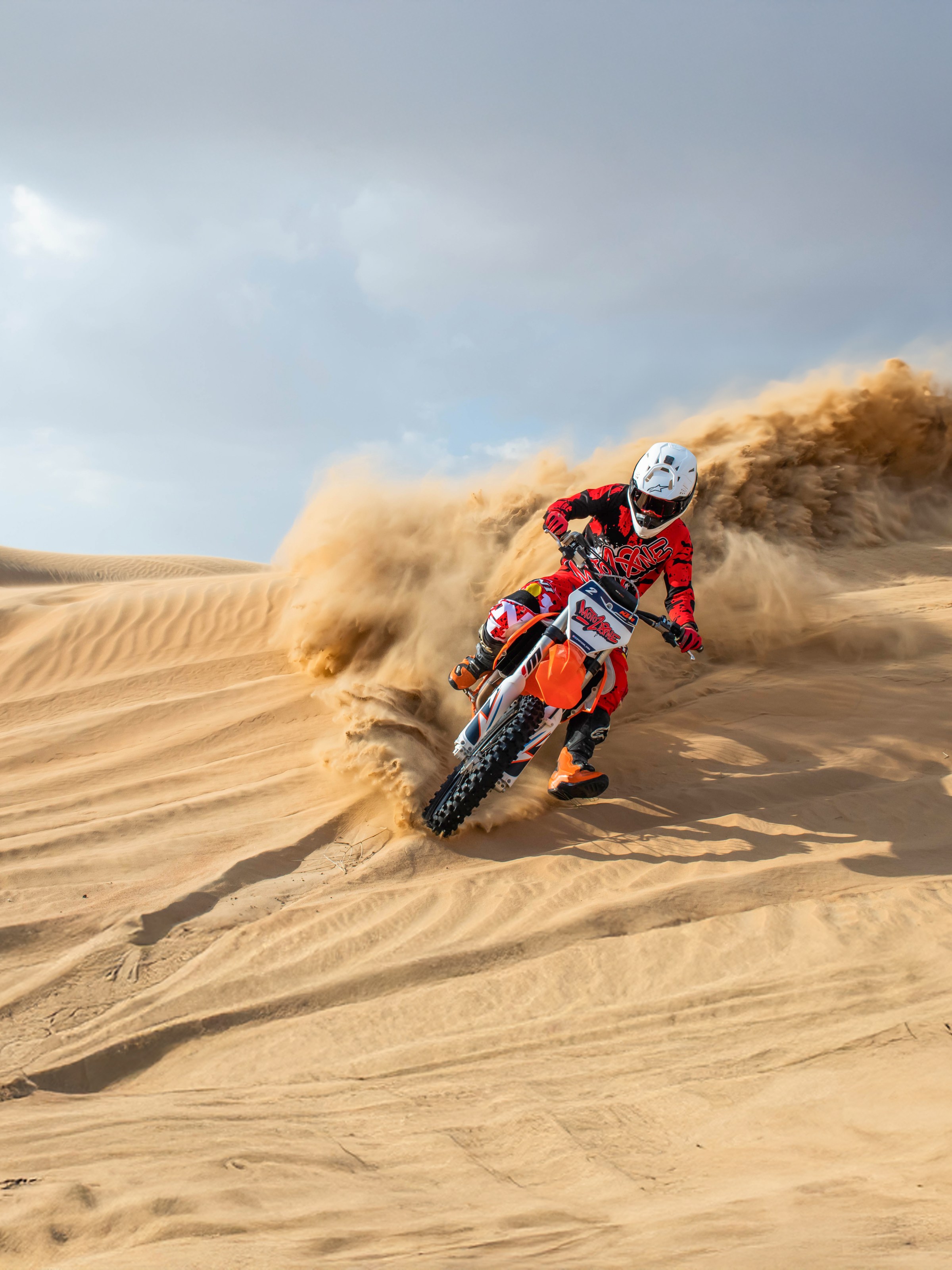 Motocross rider in red gear kicking up sand while riding on desert dunes.