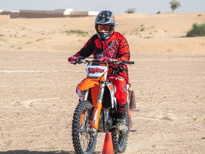 Motorcyclist in red gear rides through cones on desert terrain.