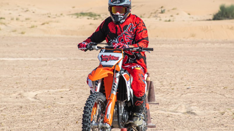 Motorcyclist in red gear rides through cones on desert terrain.