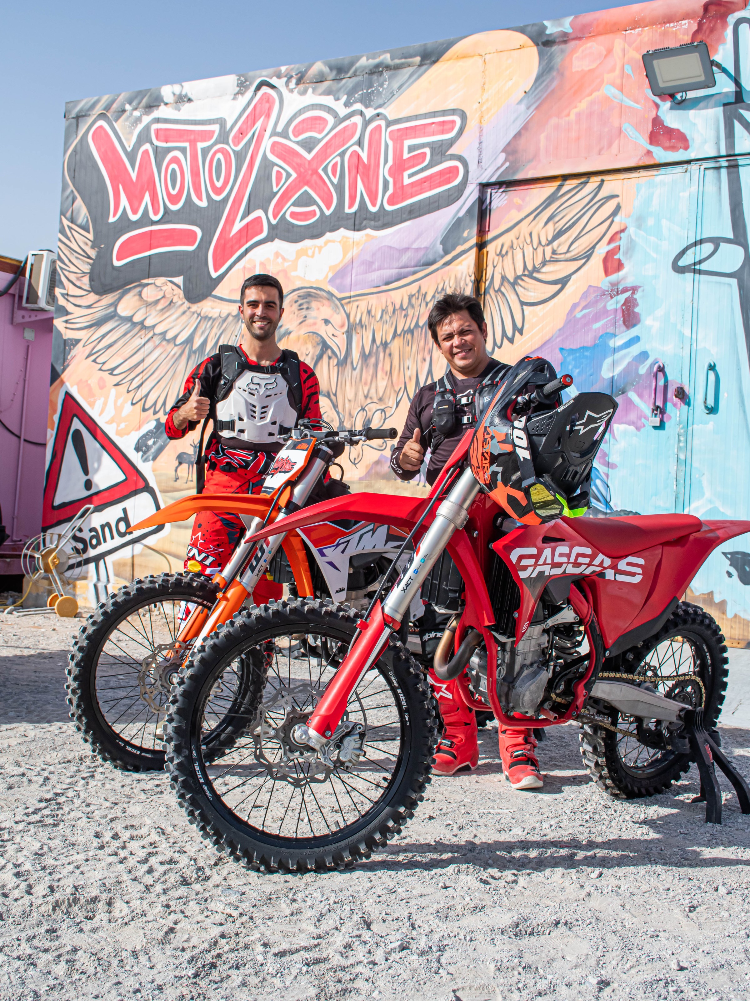 Two people in motocross gear stand beside dirt bikes, graffiti wall backdrop.