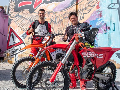Two people in motocross gear stand beside dirt bikes, graffiti wall backdrop.