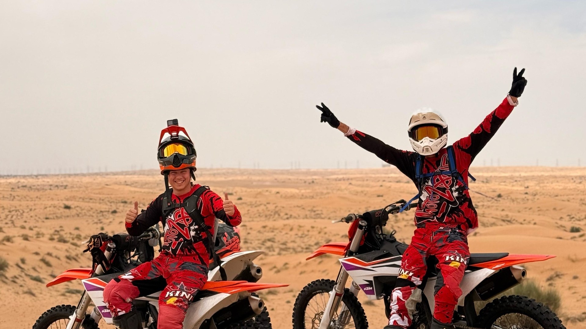 Two people on dirt bikes in the desert, wearing red gear and helmets, sandy landscape visible.