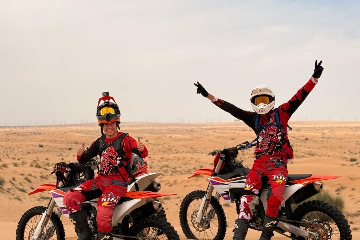 Two people on dirt bikes in the desert, wearing red gear and helmets, sandy landscape visible.