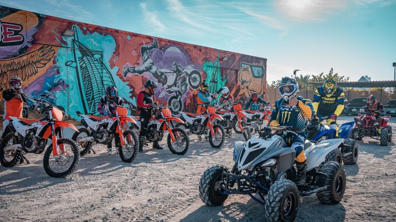 Group of riders on dirt bikes and ATVs in front of a colorful graffiti wall.