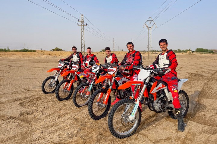 Five people in red gear on dirt bikes lined up in a desert landscape.