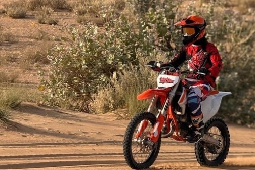 Person in red gear riding an orange dirt bike on sandy terrain.